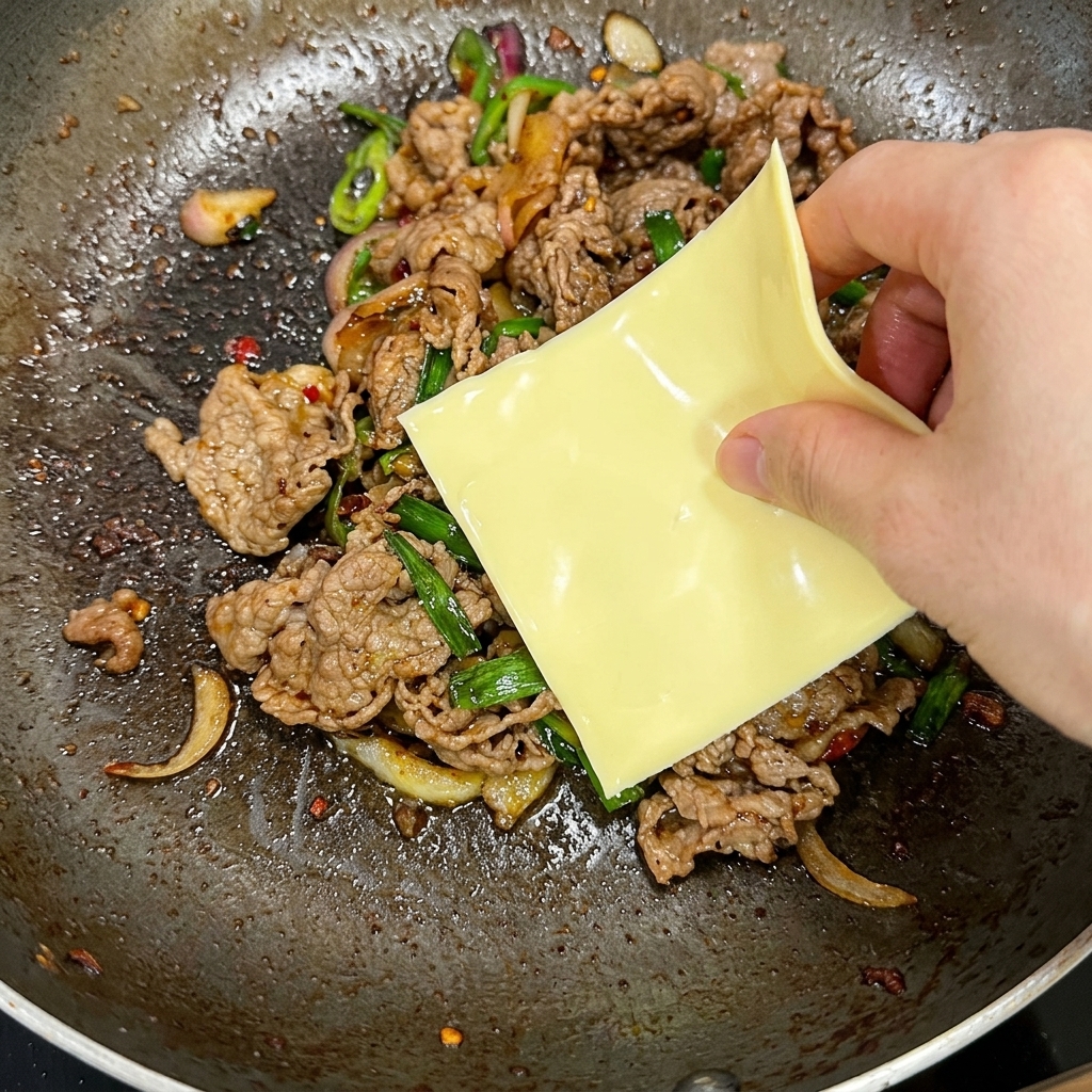 Una mano che adagia una fetta quadrata di formaggio giallo sopra il mix bollente di manzo e peperoncini in padella.