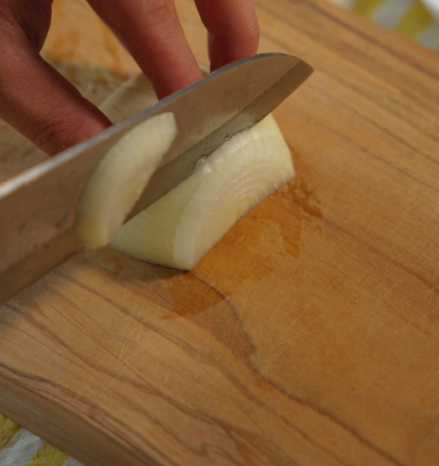 Primo piano di una mano che tiene una cipolla bianca mentre un coltello da chef affilato la taglia in pezzi uniformi su un tagliere di legno.
