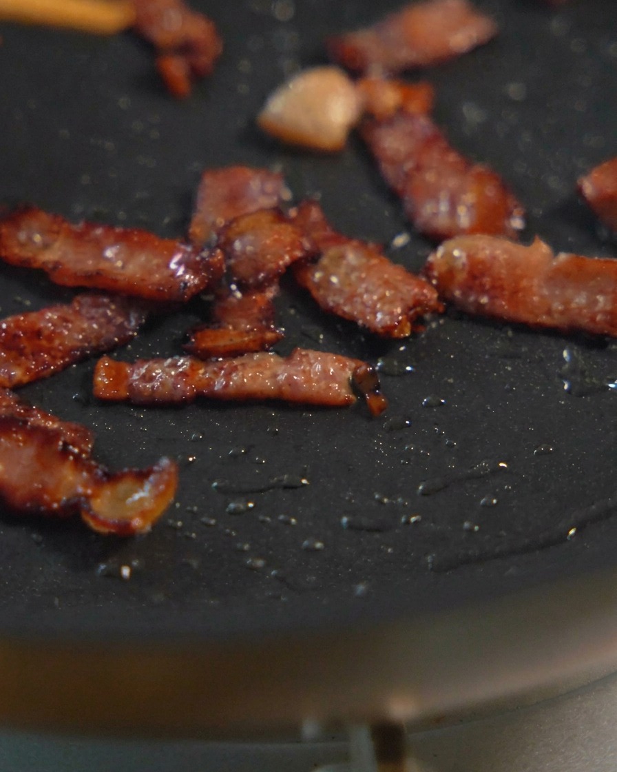 Piccoli pezzi di pancetta a dadini che sfrigolano in una padella nera, mostrando vari stadi di doratura e croccantezza.