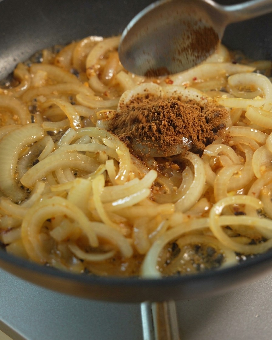 Fette di cipolla saltate in padella con un cucchiaio che aggiunge zucchero di canna e condimenti liquidi per la caramellizzazione.