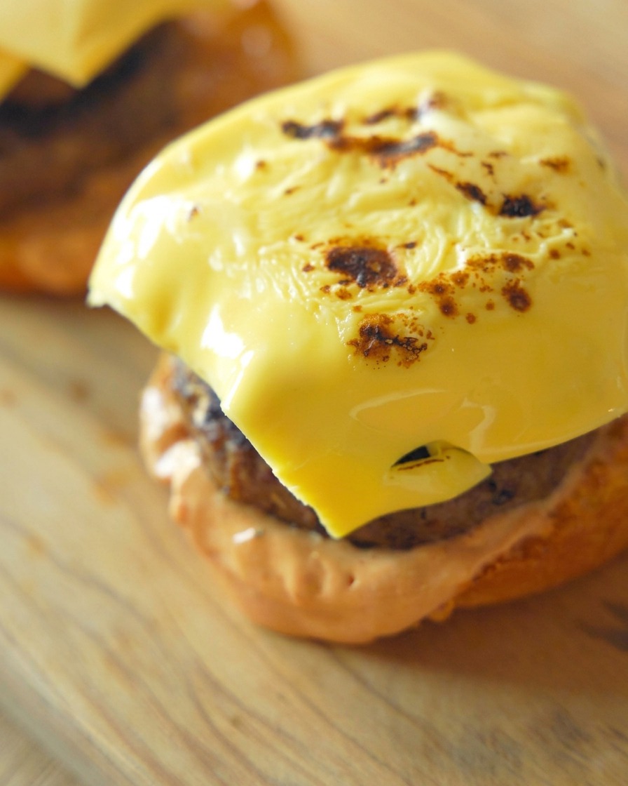 Dettaglio ravvicinato di una fetta di formaggio giallo perfettamente fusa e leggermente abbrustolita adagiata su uno spesso hamburger di manzo cotto.