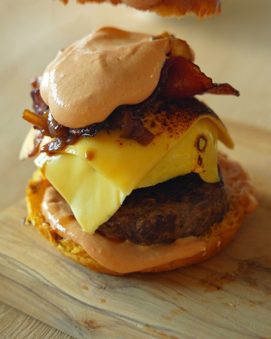 Inquadratura ravvicinata che mostra una mano che posiziona lultimo panino superiore su un alto hamburger gourmet farcito con carne, formaggio fuso, cipolle caramellate e salsa.