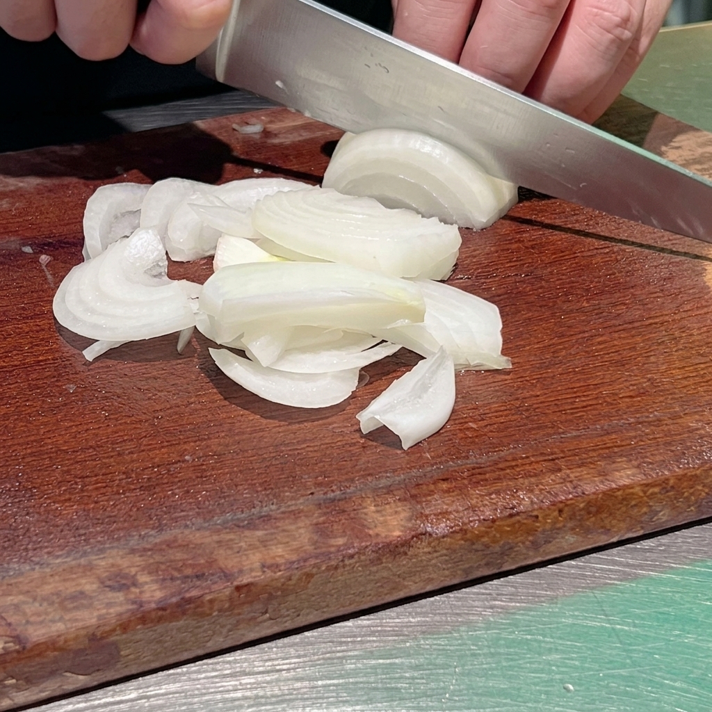 Un primo piano di uno chef che usa un coltello da cucina affilato per tagliare una cipolla bianca a strisce sottili su un tagliere di legno scuro.