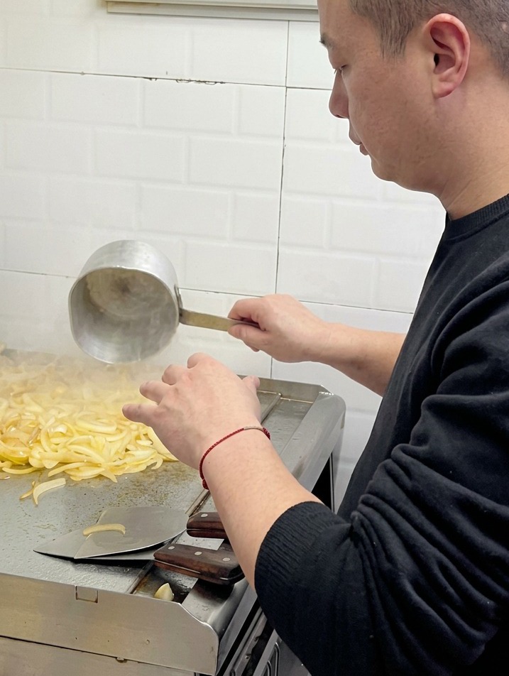 Uno chef che versa acqua da un piccolo mestolo di metallo su una pila di cipolle che si stanno ammorbidendo su una piastra professionale.