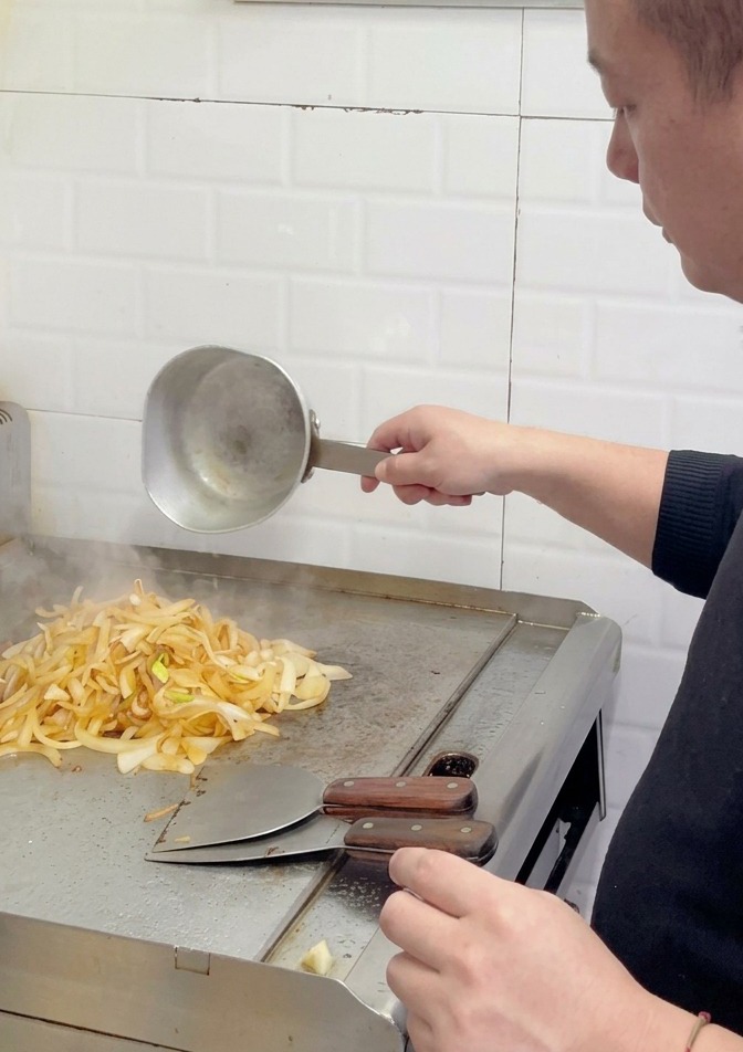 Uno chef che aggiunge altra acqua a una pila di cipolle marroncine parzialmente caramellate sulla superficie di una piastra.