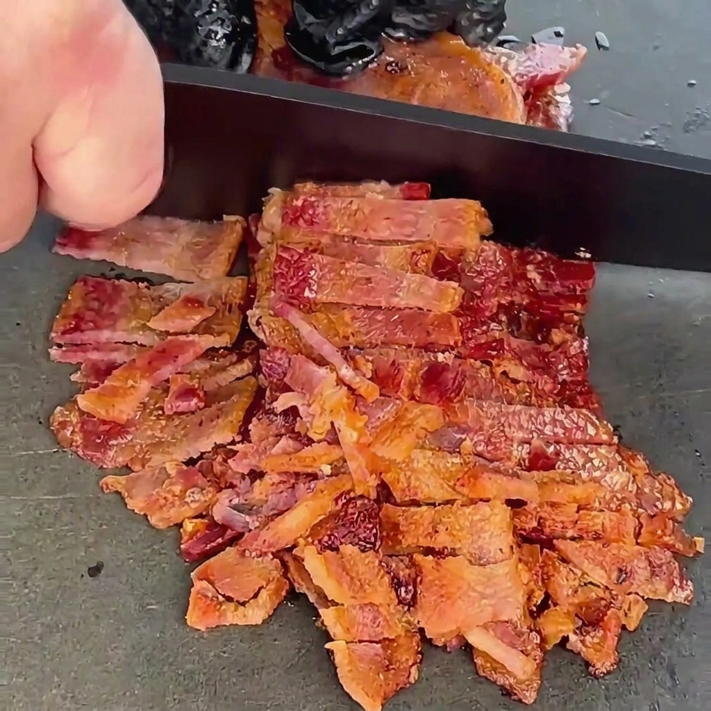 Fette di bacon croccante cotto che vengono sminuzzate finemente con un coltello da chef nero su una superficie grigia.