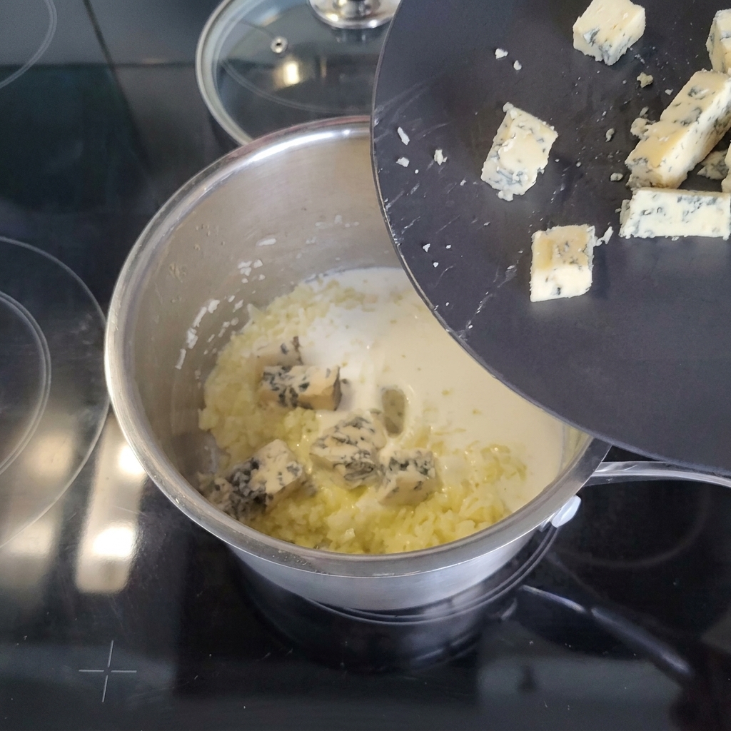 Cubetti di formaggio erborinato che scivolano da un piatto nero in un pentolino con panna.