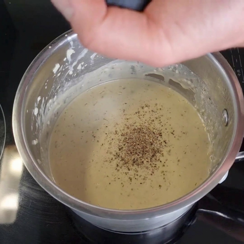 Una mano che macina pepe nero fresco da un macinino in una salsa cremosa dentro un pentolino.