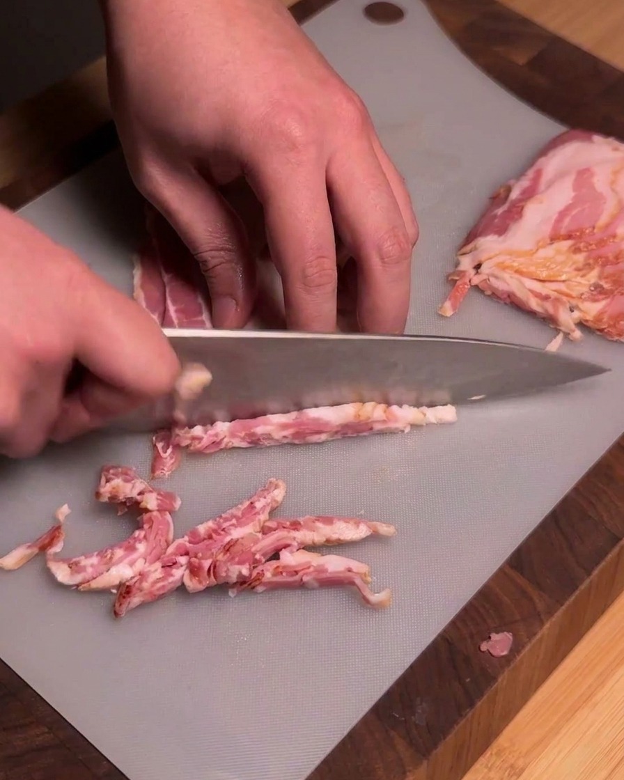 Uno chef usa un coltello da cucina affilato per tagliare strisce di bacon crudo in pezzi uniformi su un tagliere grigio.