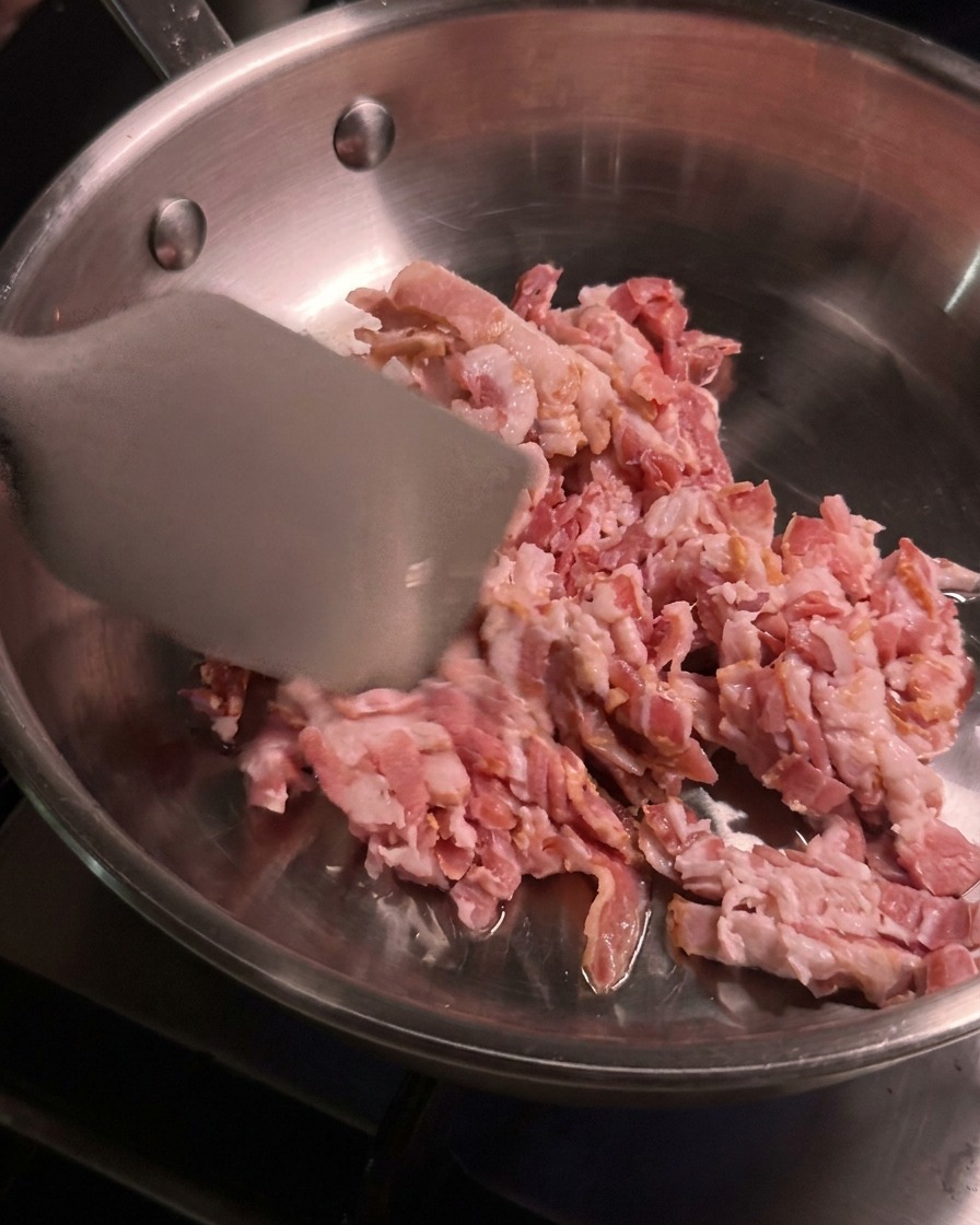 Pezzi di bacon crudo a dadini vengono mescolati e cotti con una spatola di metallo in una padella di acciaio inossidabile calda.