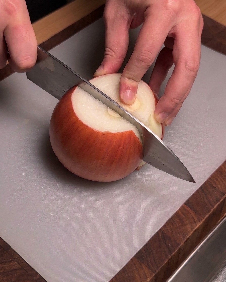 Mani che impugnano un coltello da chef per tagliare una cipolla dorata sbucciata su un tagliere grigio.