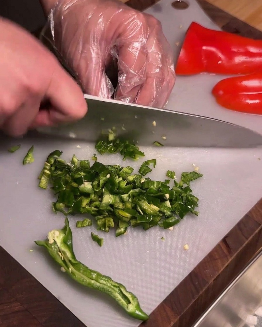 Una persona con guanti di plastica trasparenti trita finemente peperoncini verdi grinzosi privati dei semi su un tagliere.