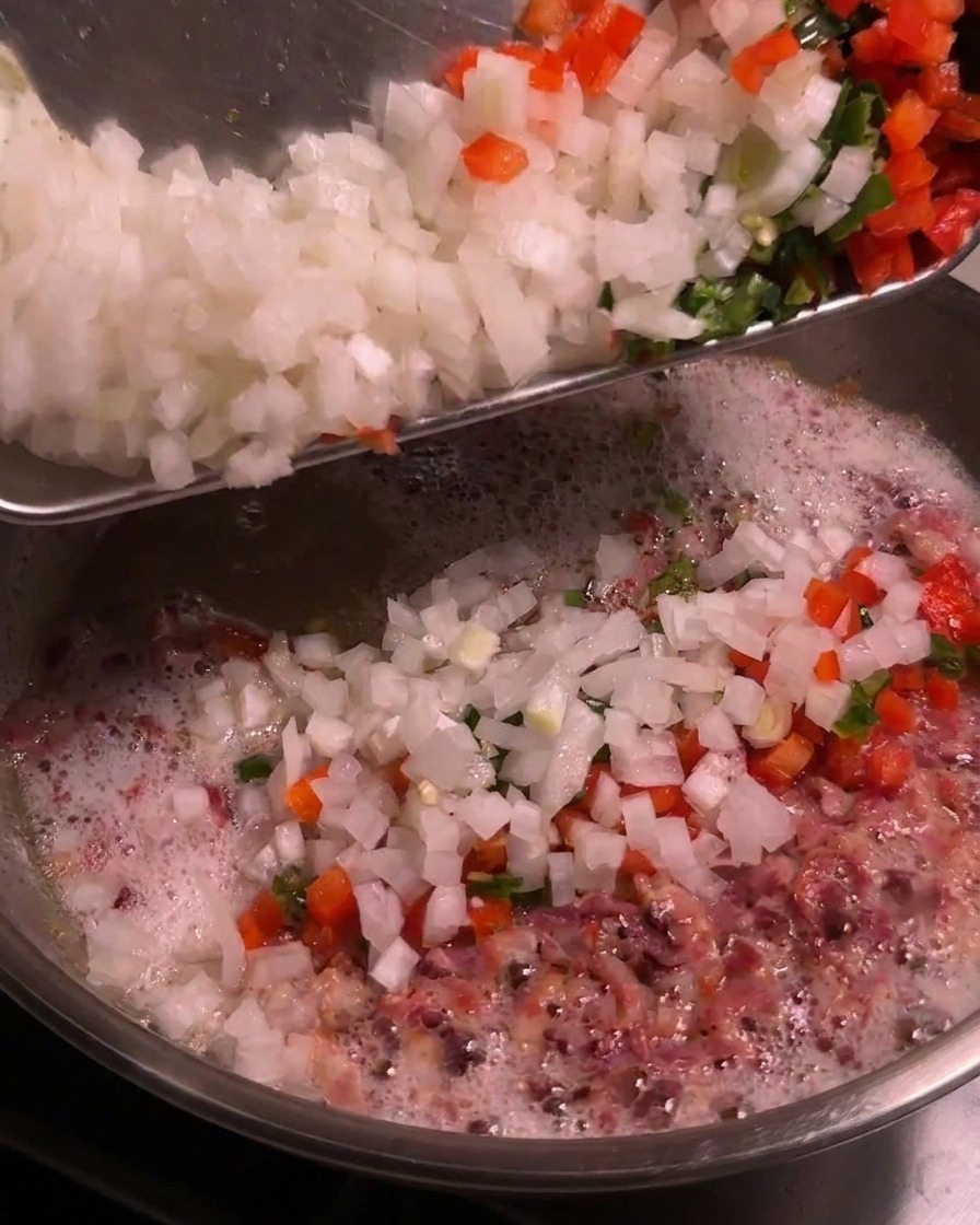 Un vassoio con cipolle bianche a dadini e peperoncini colorati tritati viene versato in una padella sfrigolante con il grasso del bacon.