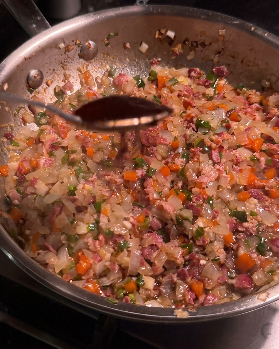 Un cucchiaio di salsa chipotle scura sospeso sopra una padella piena del mix di bacon e verdure.