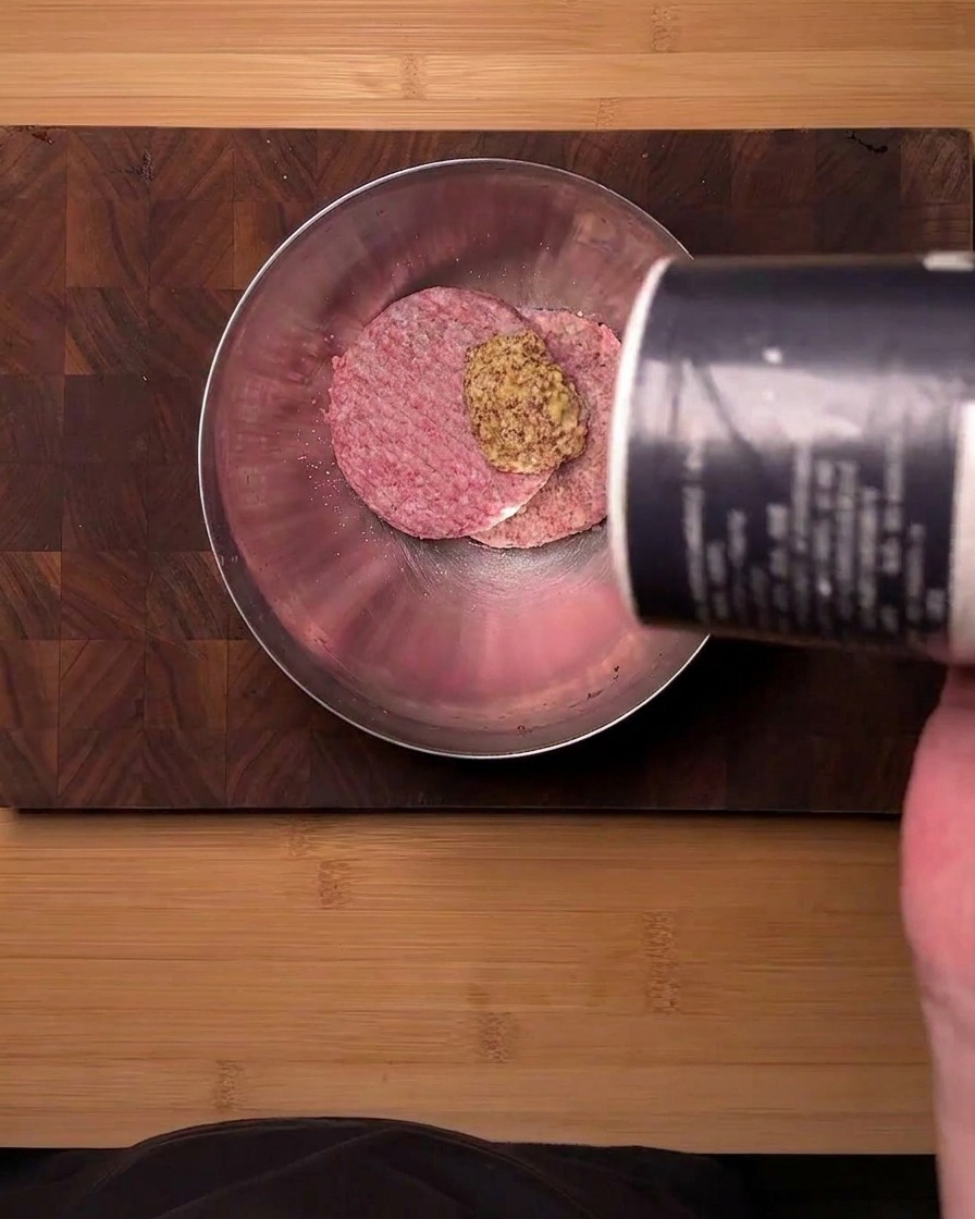 Vista dallalto di carne macinata di manzo cruda in una ciotola dacciaio condita con senape in polvere e sale versato da un contenitore.