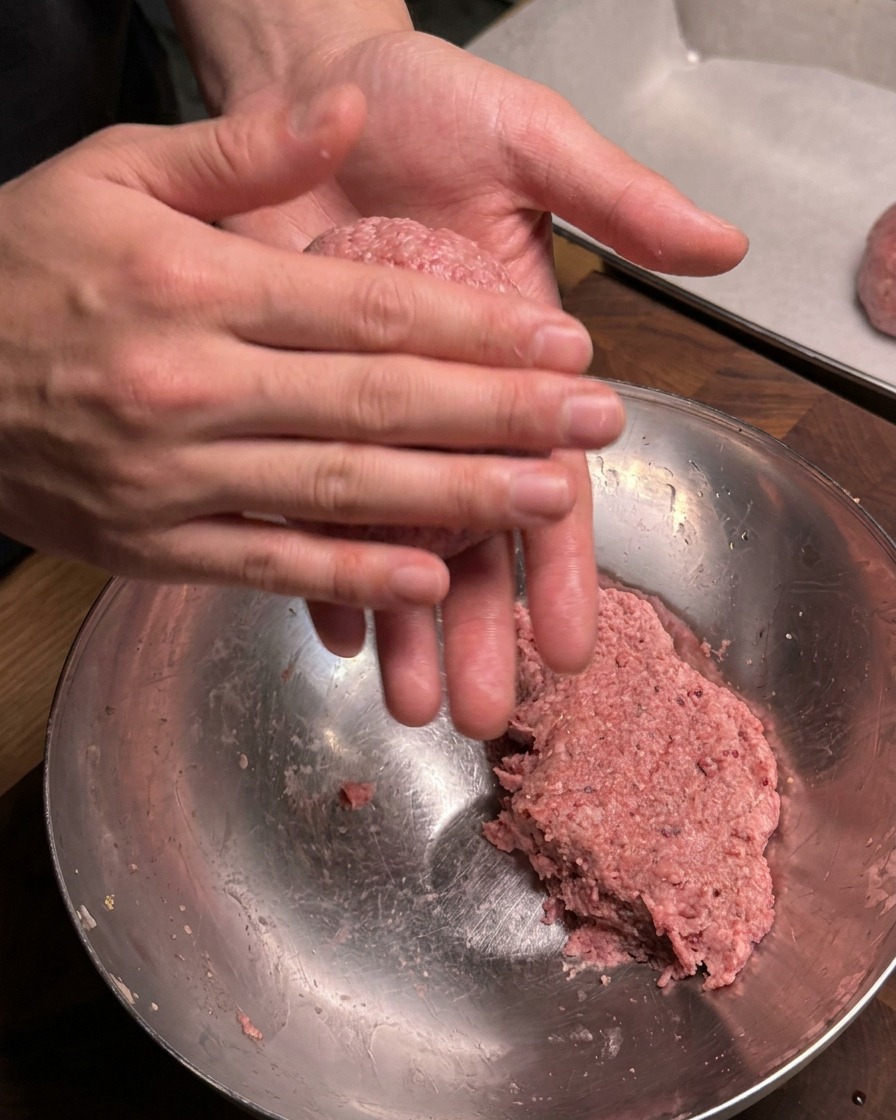 Primo piano di mani che arrotolano e formano con decisione una porzione di carne macinata condita in una polpetta rotonda sopra una ciotola di metallo.