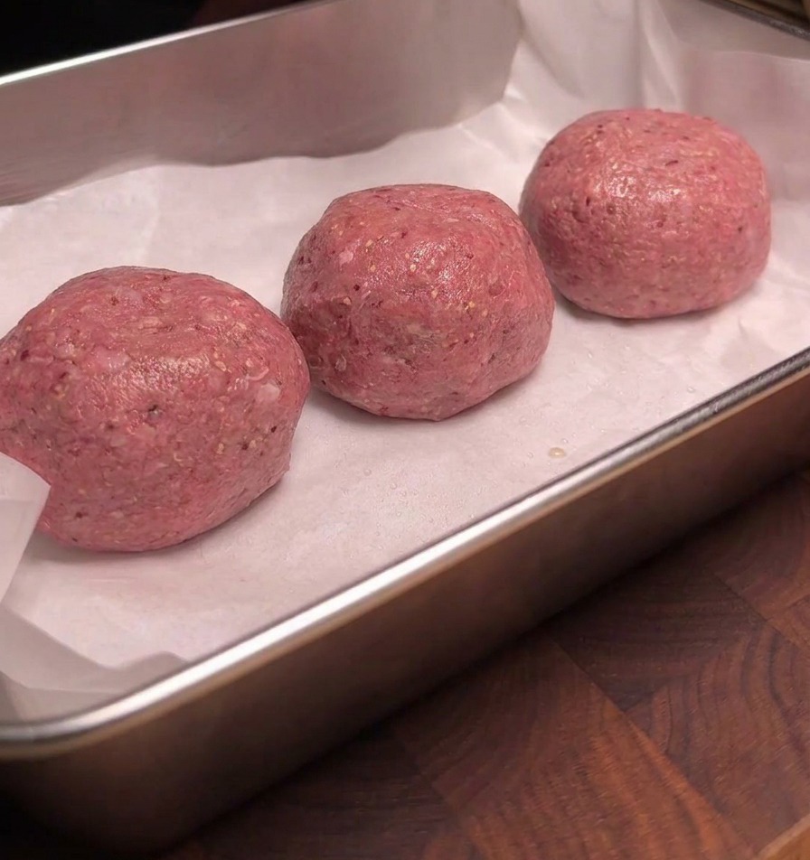 Tre polpette di manzo crudo disposte ordinatamente su carta da forno bianca in un vassoio di metallo, con le superfici lucide e uniformemente bagnate di fumo liquido.