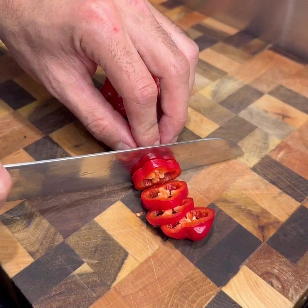 Una persona usa un coltello da cucina per tagliare a rondelle dei peperoncini rossi freschi su un tagliere di legno.