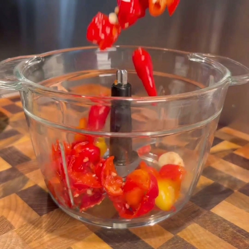 Peperoncini rossi fermentati e spicchi daglio scolati che cadono nella ciotola trasparente di un robot da cucina.
