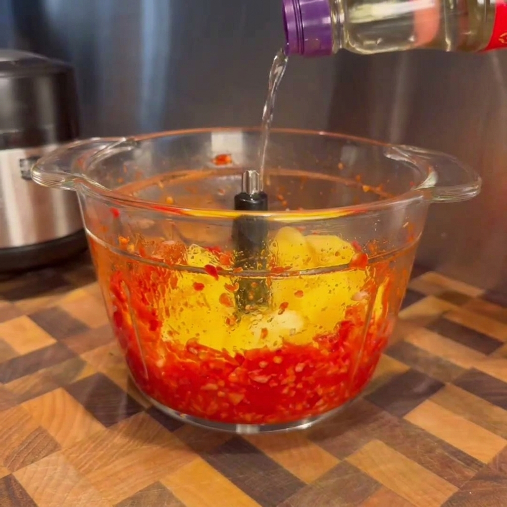 Liquido trasparente versato da una bottiglia nella ciotola di vetro di un robot da cucina contenente peperoncini rossi frullati e pezzi di mango giallo brillante.