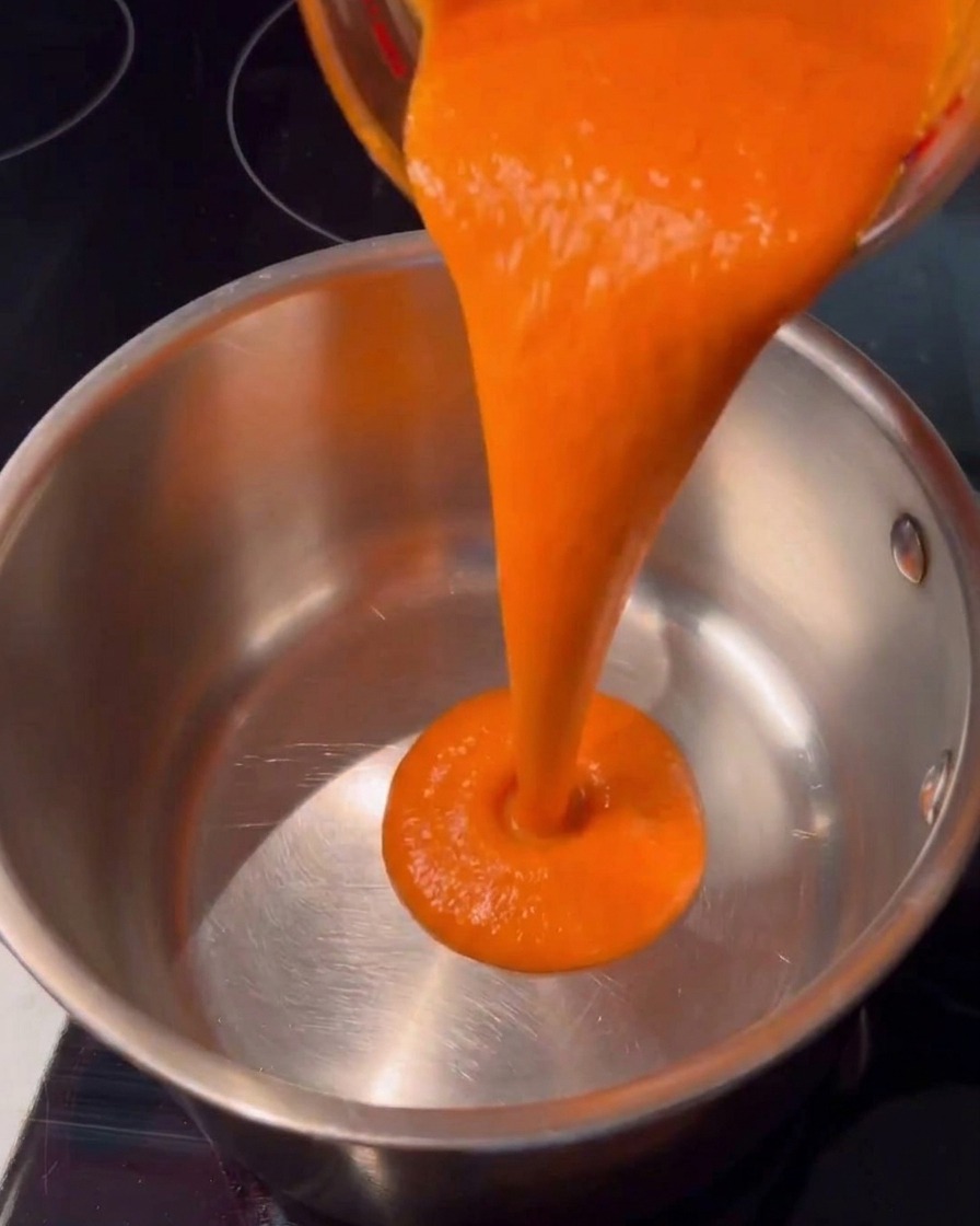 Salsa piccante arancione brillante che viene versata dolcemente da una tazza di vetro in una casseruola in acciaio inox pulita su un piano cottura scuro.