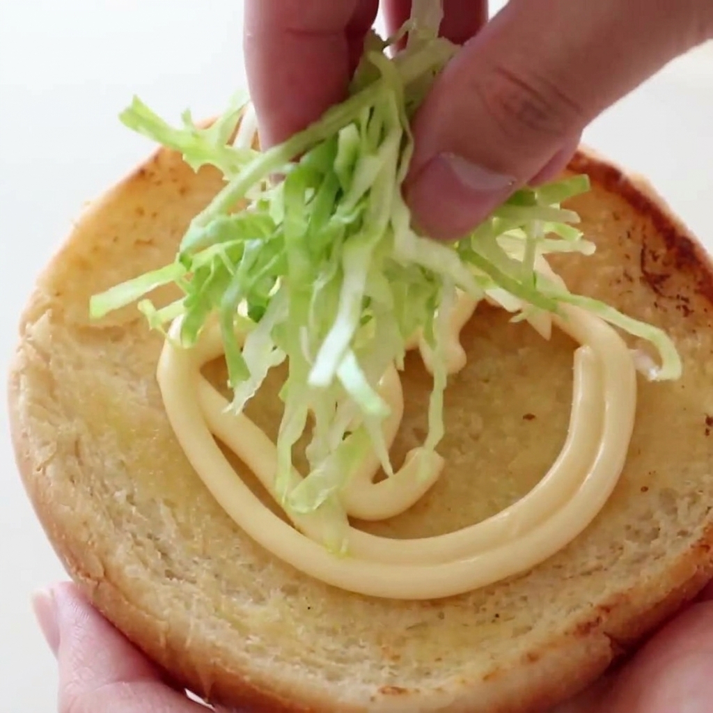 Una mano che posiziona una manciata di lattuga verde croccante e finemente tagliata su un panino tostato stratificato con una spirale di maionese cremosa.