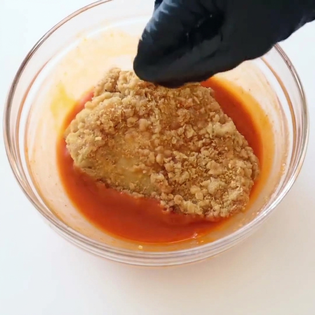 Una mano che indossa un guanto nero mentre immerge un filetto di pollo fritto croccante e dorato in una ciotola trasparente piena di salsa buffalo rosso vivo.