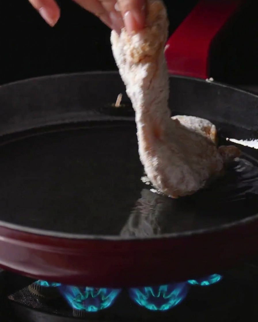 Una mano che adagia una sovracoscia di pollo infarinata in una padella con olio caldo su un fornello a gas acceso.