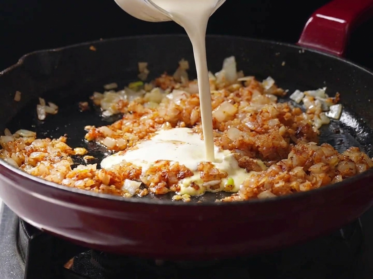 Panna liquida versata da un contenitore di vetro in una padella con gli odori soffritti e conditi.
