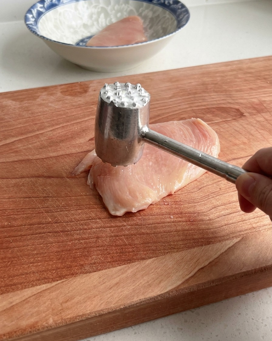 Una mano che usa un batticarne in metallo per battere delicatamente un petto di pollo crudo su un grande tagliere di legno.