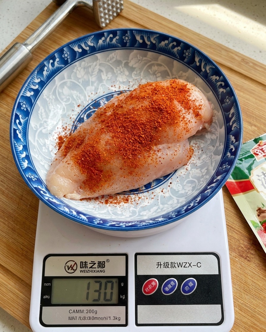 Un petto di pollo crudo in una ciotola di ceramica decorata posizionata su una bilancia digitale da cucina, cosparso con abbondante polvere speziata arancione-rossastra.