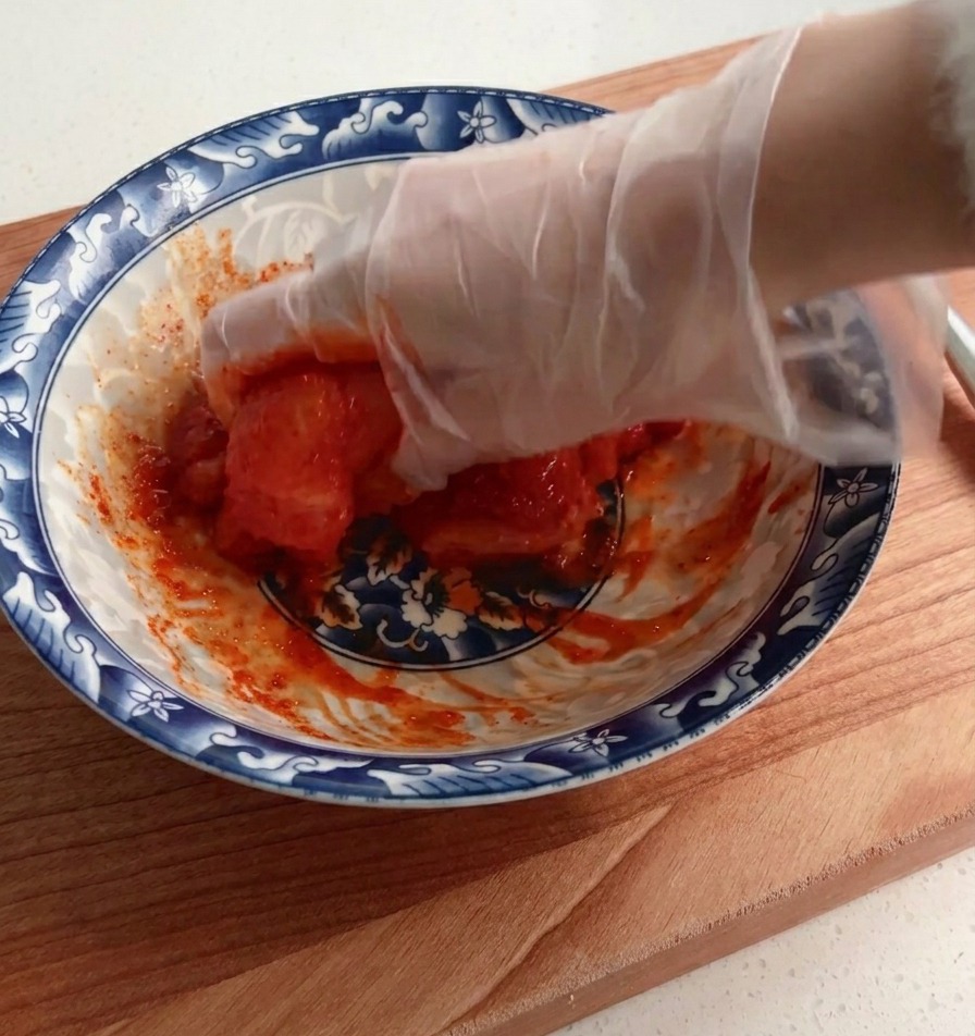 Una mano con un guanto che distribuisce e mescola accuratamente un petto di pollo crudo in una densa marinatura rossa allinterno di una ciotola decorata.