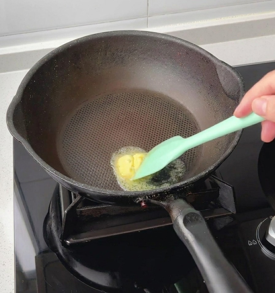 Una spatola in silicone verde che scioglie un cubetto di burro giallo al centro di una padella nera calda.