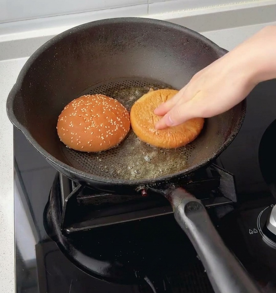 Una mano che preme le due metà di un panino al sesamo rivolte verso il basso nel burro fuso allinterno di una padella.