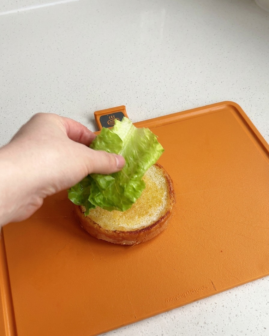 Una mano che posiziona una foglia di insalata verde fresca e croccante sopra la base dorata e tostata di un panino, su un tagliere arancione.