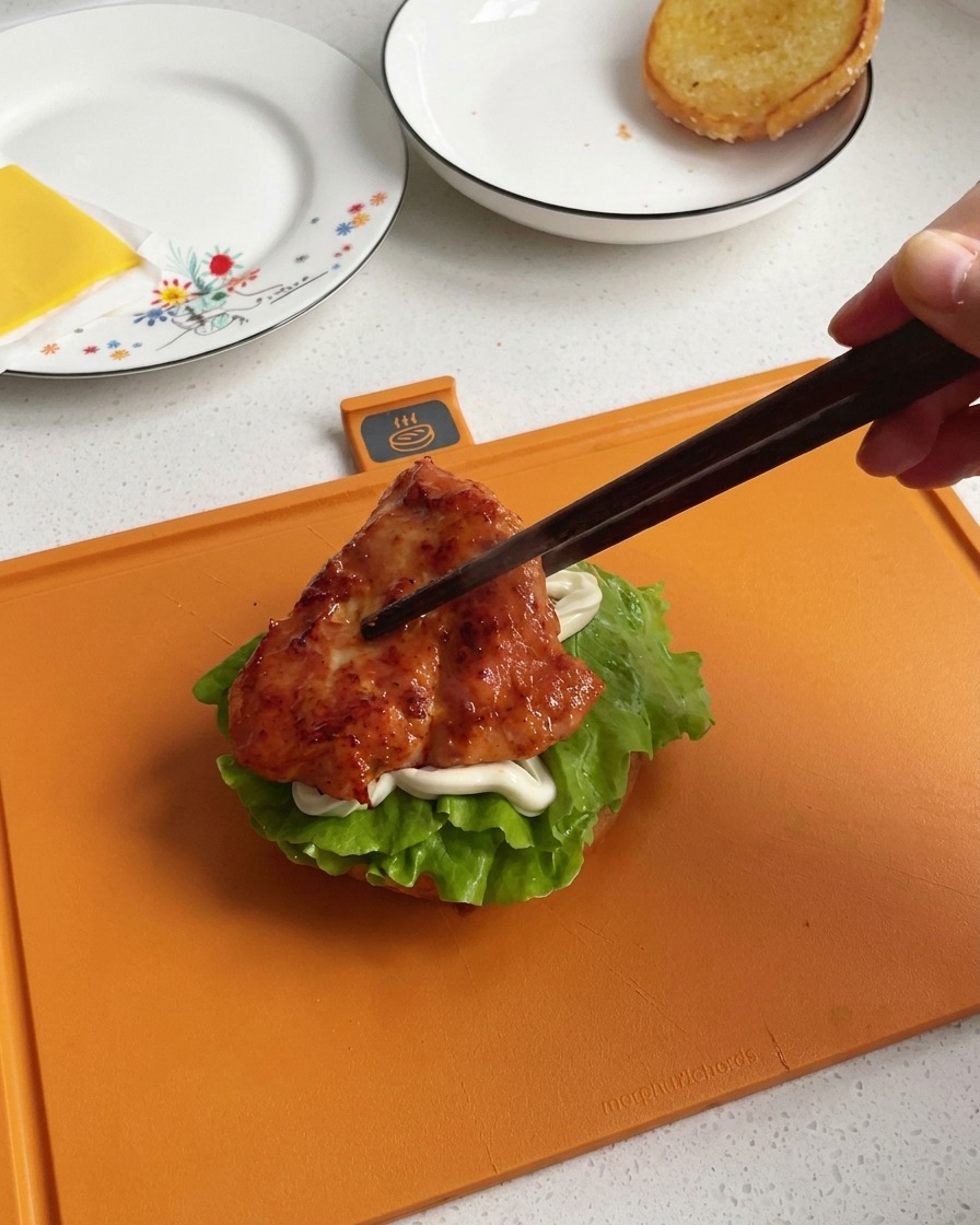 Una mano che usa delle bacchette di legno scuro per trasferire un pezzo di pollo cotto in padella, lucido e ben caramellato, sopra la base del burger.
