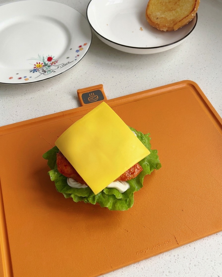 Una fetta quadrata di formaggio fuso giallo brillante adagiata sopra un pezzo di pollo appena cotto in un burger parzialmente assemblato.
