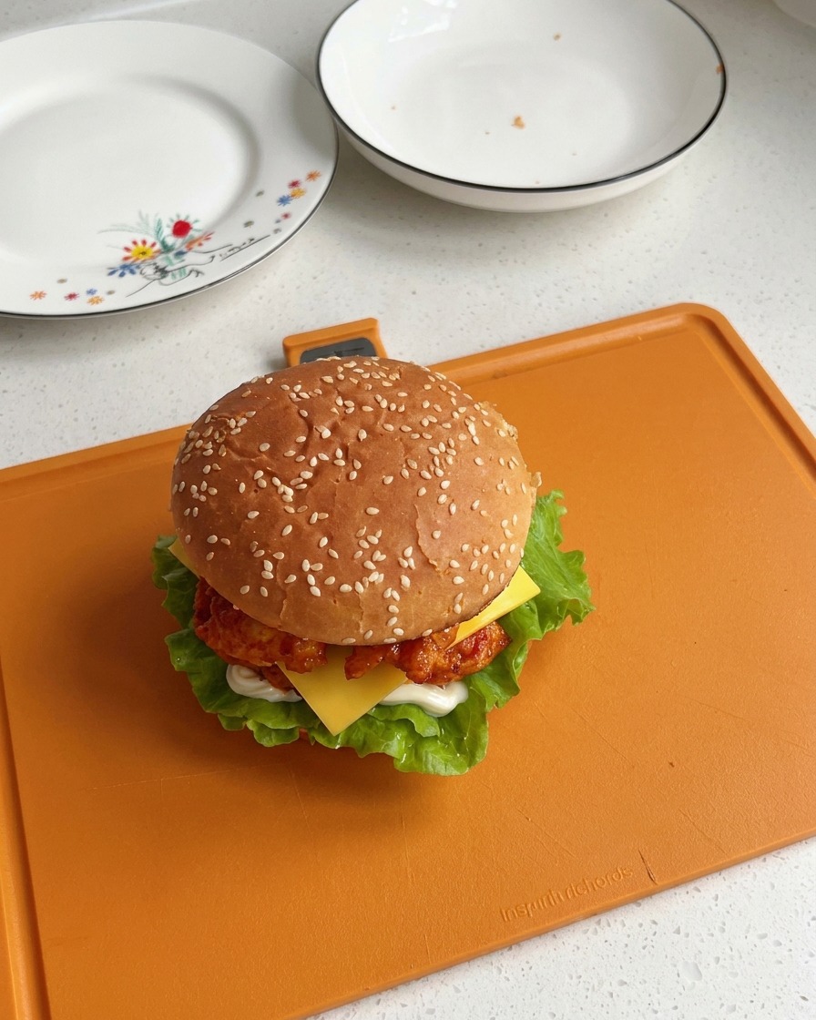 Un burger di pollo Orleans completamente assemblato e chiuso con il panino al sesamo, che mostra strati visibili di insalata, pollo e formaggio fuso.