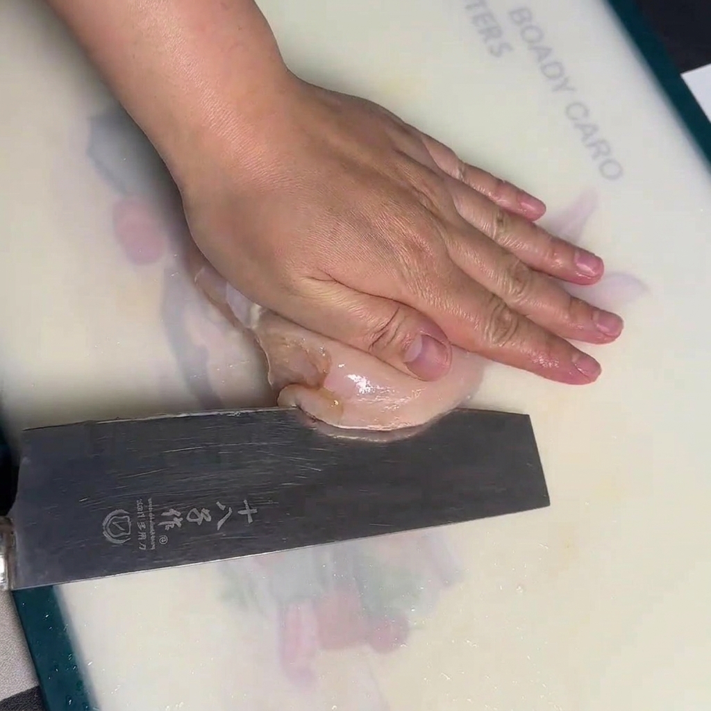 Una mano tiene fermo un petto di pollo crudo su un tagliere bianco mentre lo taglia con cura in senso orizzontale con un grosso coltello da chef.