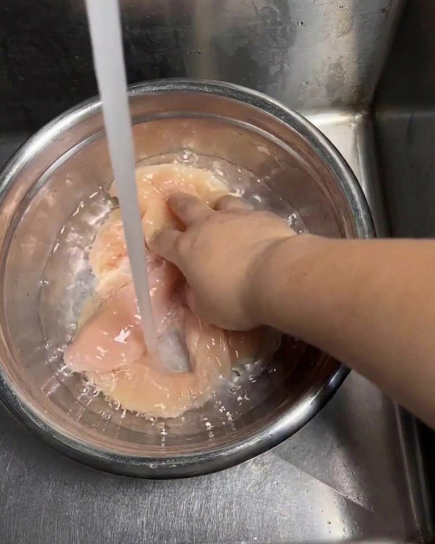 Un getto di acqua fresca del rubinetto riempie una ciotola di acciaio contenente le cotolette di pollo crudo mentre una mano muove i pezzi per sciacquarli.