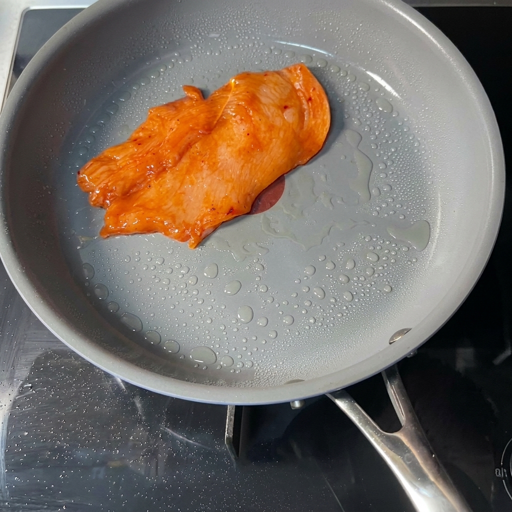 Una singola cotoletta di pollo marinata di colore arancione cuoce in una padella antiaderente grigia con visibili goccioline dolio in superficie.