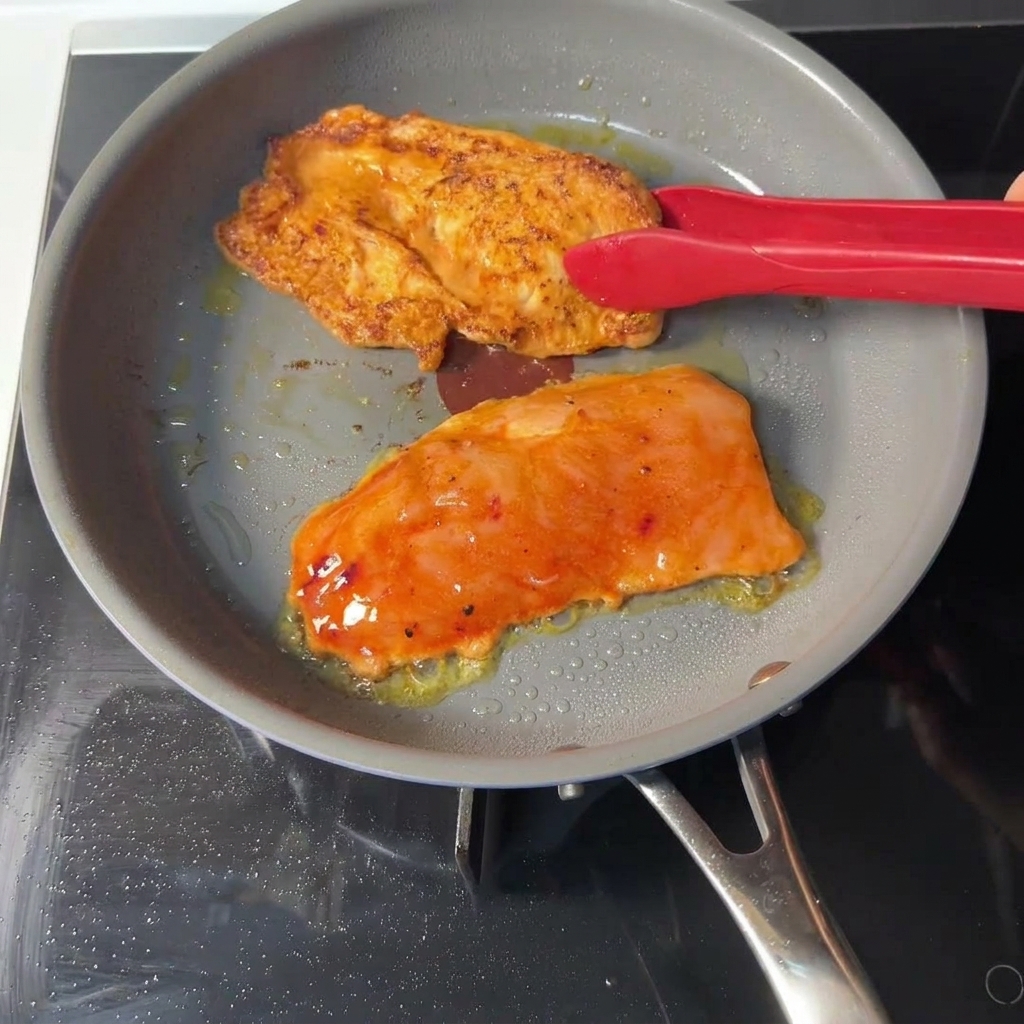 Delle pinze rosse girano una cotoletta di pollo arancione in una padella grigia, mostrando una crosticina dorata e rosolata sul lato già cotto.
