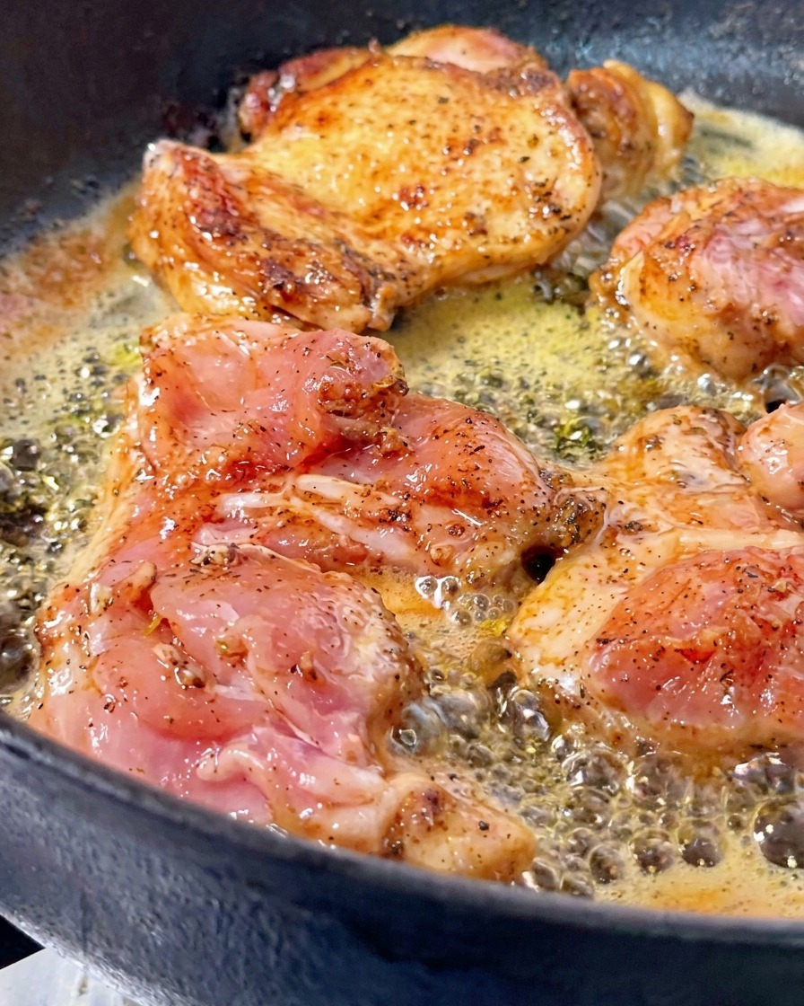 Cosce di pollo cotte nel burro spumeggiante in una padella di ghisa, con un pezzo girato che mostra una crosticina dorata e croccante.