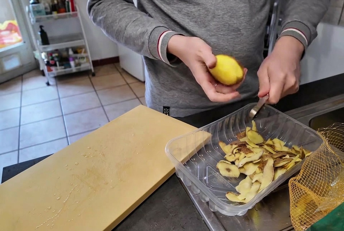 Una persona che pela una patata gialla cruda con un coltellino sopra un contenitore di plastica trasparente contenente le bucce.