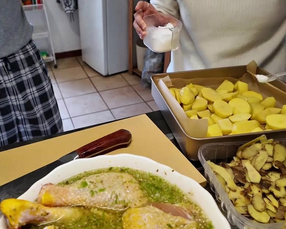 Pezzi di patate crude disposti su una teglia foderata di carta forno mentre vengono salati spargendo il sale da un contenitore trasparente.