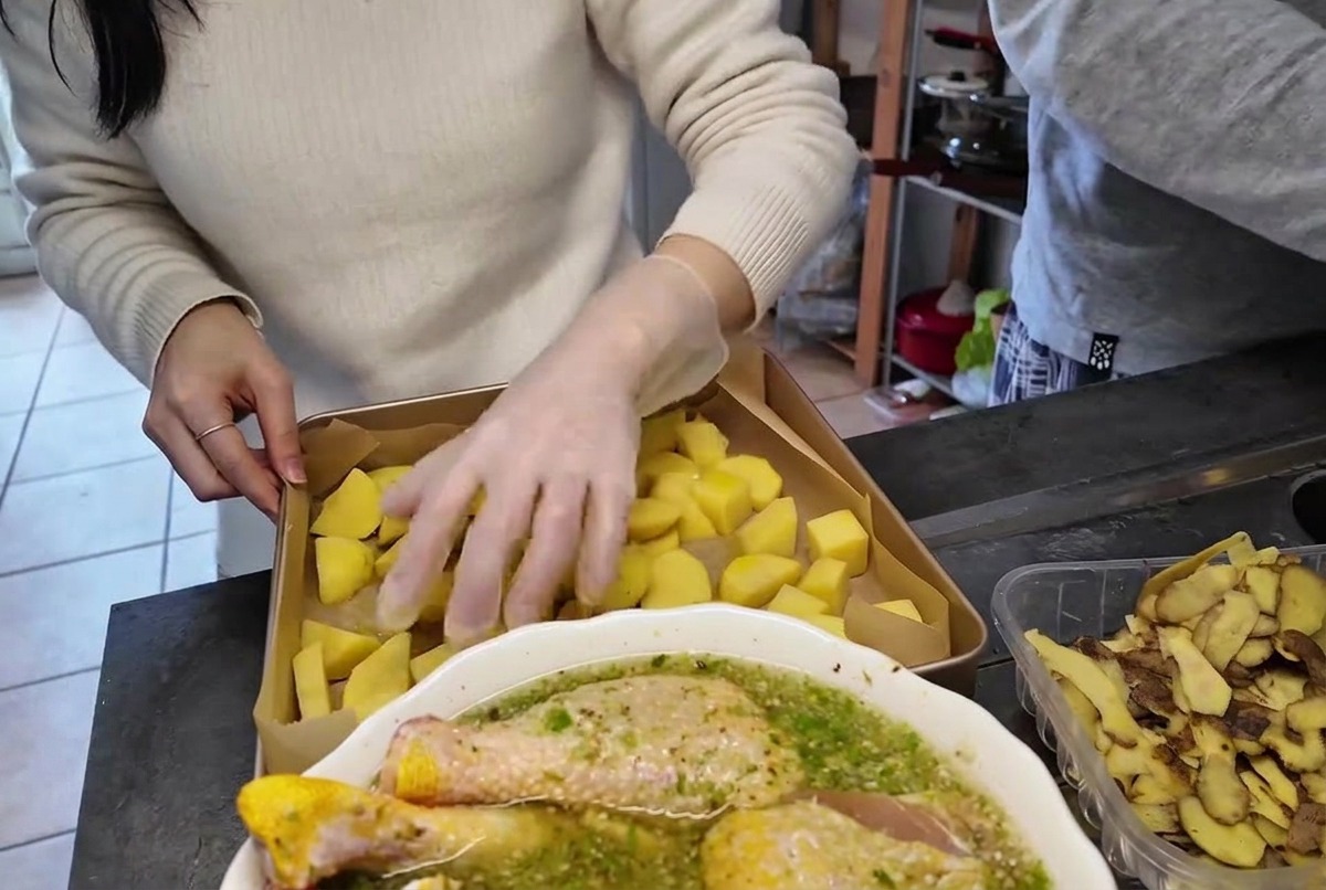 Una mano con un guanto di plastica trasparente che gira e mescola i pezzi di patate in una teglia di metallo.