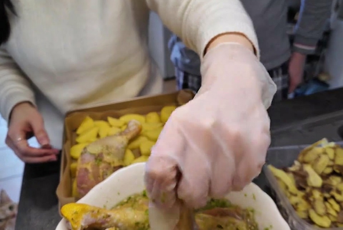 Una mano con il guanto che solleva un fuso di pollo crudo ricoperto di marinata verde alle erbe da una ciotola bianca.