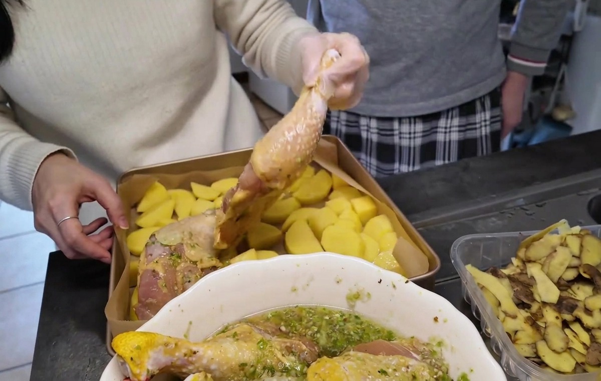 Cosce di pollo crude marinate disposte con la pelle verso lalto sopra uno strato di patate a pezzi in una teglia dorata.