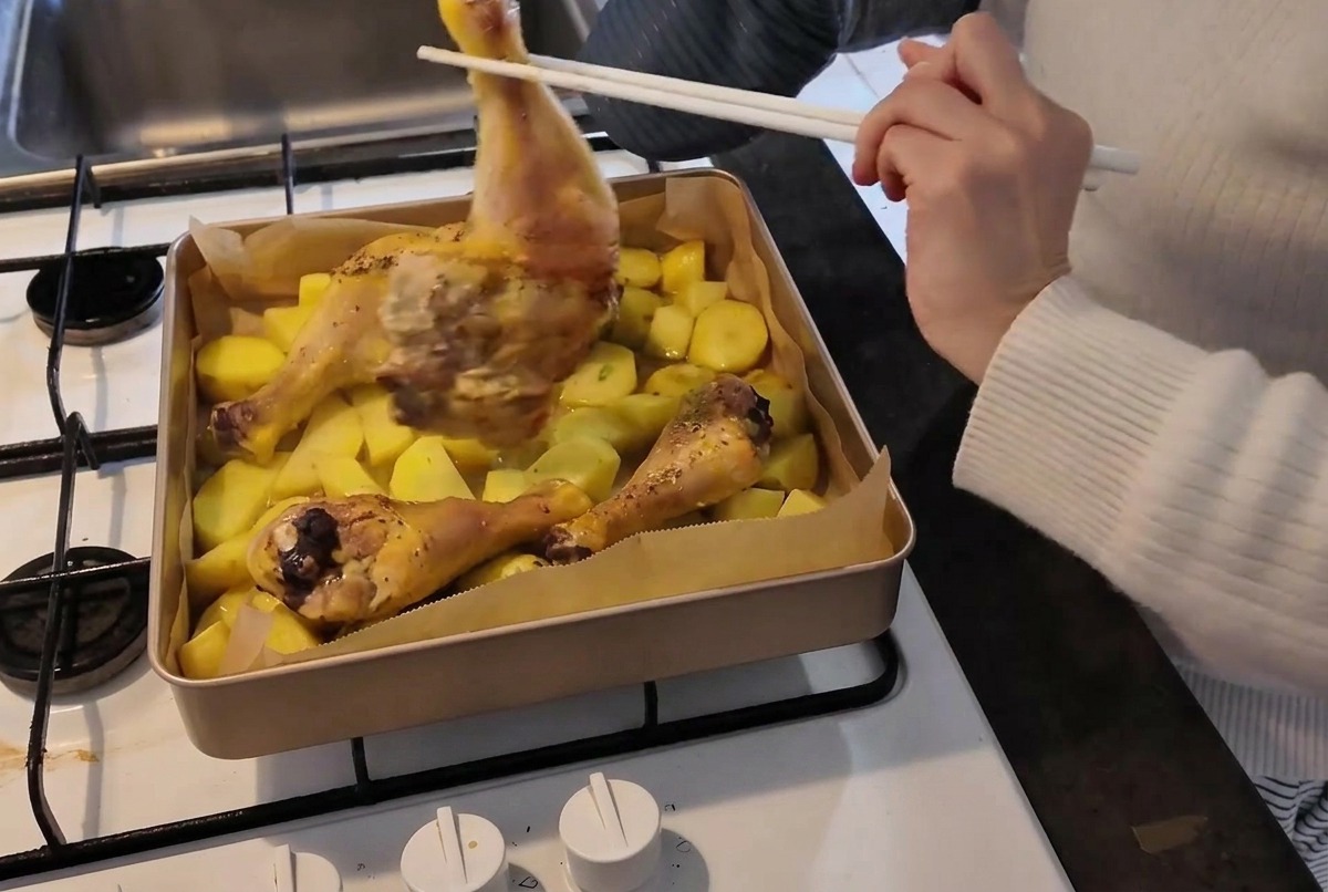 Una persona con un guanto da forno usa le bacchette per girare una coscia di pollo parzialmente cotta in una teglia bollente piena di patate.