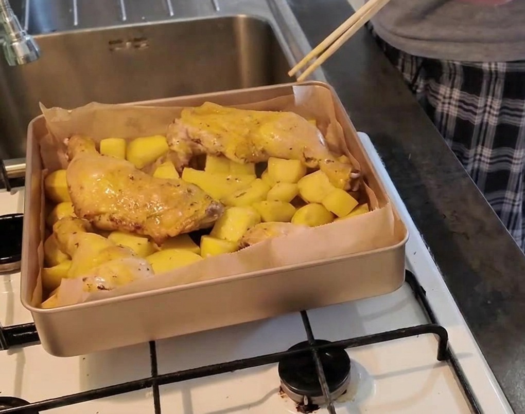 Una persona usa un paio di bacchette per pungere la carne di una coscia di pollo adagiata su un letto di patate per verificarne la cottura.